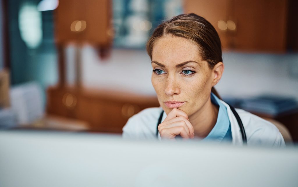 Eine Ärztin im weißen Kittel und mit Stethoskop ist nachdenklich über einem Computermonitor zu sehen, mit einer Hand am Kinn. Sie wirkt rätselnd, während sie sich auf die Diagnose von Histaminintoleranz konzentriert. Der Ausdruck ihrer Miene vermittelt die gründliche und komplexe Natur der Diagnose dieses Krankheitsbildes.