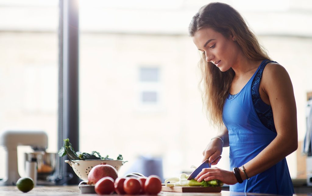 Eine Frau mit blauer Küchenschürze schneidet Lauch mit einem Messer auf einem Schneidebrett. Neben ihr sind Granatapfel, Tomaten und eine Schüssel mit grünem Gemüse zu sehen. Der Hintergrund deutet auf eine gesunde und ausgewogene Ernährung hin, die im Zusammenhang mit der Behandlung einer Histaminintoleranz steht.
