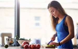 Eine Frau mit blauer Küchenschürze schneidet Lauch mit einem Messer auf einem Schneidebrett. Neben ihr sind Granatapfel, Tomaten und eine Schüssel mit grünem Gemüse zu sehen. Der Hintergrund deutet auf eine gesunde und ausgewogene Ernährung hin, die im Zusammenhang mit der Behandlung einer Histaminintoleranz steht.