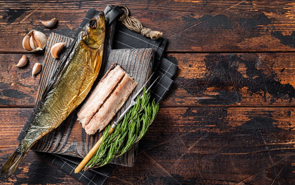 Ein ganzer Fisch und ein Fischfilet liegen auf einem Holz-Schneidebrett, begleitet von einer Fischgabel und einem Rosmarinzweig. Fisch ist ein Lebensmittel, das oft viel Histamin enthält, besonders bei bestimmten Sorten wie Hering, Makrele oder Thunfisch, da Mikroorganismen während der Lagerung oder Verarbeitung Histamin produzieren können.