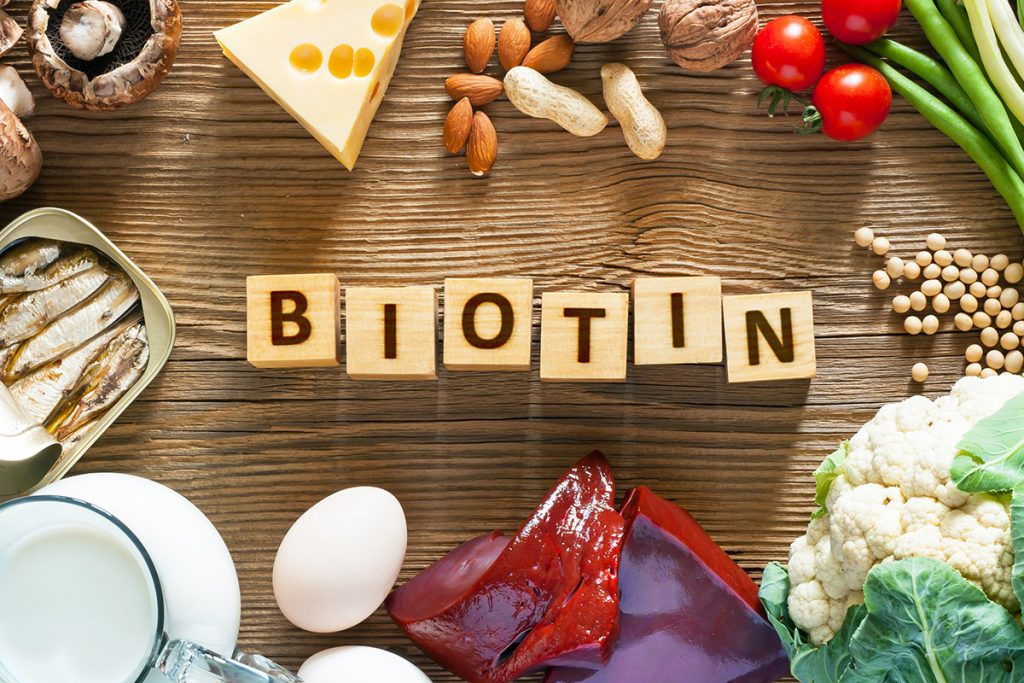 Das Bild zeigt eine Auswahl an biotinreichen Lebensmitteln wie Sojabohnen, Hühnereier, Rinderleber und Fisch, die attraktiv arrangiert sind. In der Mitte des Bildes befindet sich eine Tafel mit der Aufschrift "Biotin". Diese Lebensmittel sind bekannt dafür, Biotin zu enthalten, ein wasserlösliches B-Vitamin, das eine wichtige Rolle für die Hautgesundheit, den Stoffwechsel und das Haarwachstum spielt. Auf dem Bild wird die Bedeutung von Biotin visuell unterstrichen, mit einer klaren Betonung auf natürlichen Quellen dieses Vitamins.