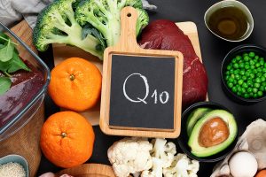 Appetitlich angerichtete Lebensmittel mit hohem Gehalt an Coenzym Q10, darunter Orangen, Leber, Brokkoli, Erbsen, Eier und pflanzliche Öle. In der Bildmitte steht eine Kreidetafel mit der Aufschrift „Q10“. Die Darstellung verweist auf die Bedeutung von Coenzym Q10 als vitaminähnliche Substanz, die vor allem in tierischen Produkten sowie ausgewählten pflanzlichen Lebensmitteln enthalten ist und eine zentrale Rolle in der Energieproduktion der Mitochondrien spielt.