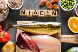 Das Bild zeigt eine Auswahl an Niacin-reichen Lebensmitteln, darunter Sardellen, Schweinefleisch und Schweineleber, die auf einem Tisch arrangiert sind. In der Mitte des Bildes befindet sich eine Tafel mit der Aufschrift "Niacin". Diese Lebensmittel sind hervorragende Quellen für Niacin (Vitamin B3), das eine wichtige Rolle für die Gesundheit der Schleimhäute spielt und im Stoffwechsel von Nährstoffen unerlässlich ist.