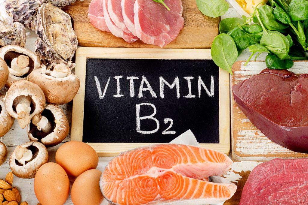 Das Bild zeigt eine Vielzahl von Vitamin B2-reichen Lebensmitteln wie Nüsse, Schweinefleisch, Schweineleber, Spinat, Käse, Eier und Lachs, die ansprechend auf einem Tisch arrangiert sind. In der Mitte des Bildes befindet sich eine Tafel mit der Aufschrift "Vitamin B2". Diese Lebensmittel sind ausgezeichnete Quellen für Riboflavin, das eine wichtige Rolle für die Gesundheit der Haut und Schleimhäute spielt.