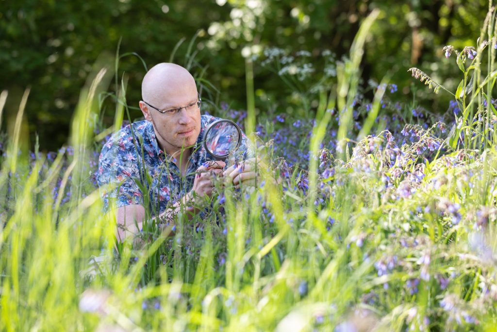 Ernährungswissenschaftler Dr. Steffen Jakobs begutachtet auf einer Wiese mit einer Lupe Pflanzen mit lila-farbenen Blüten. Das Bild unterstreicht seine stetige Arbeit an der kontinuierlichen Verbesserung von Rezepturen, wie sie auf der VitaPart®-Philosophie-Seite beschrieben wird.