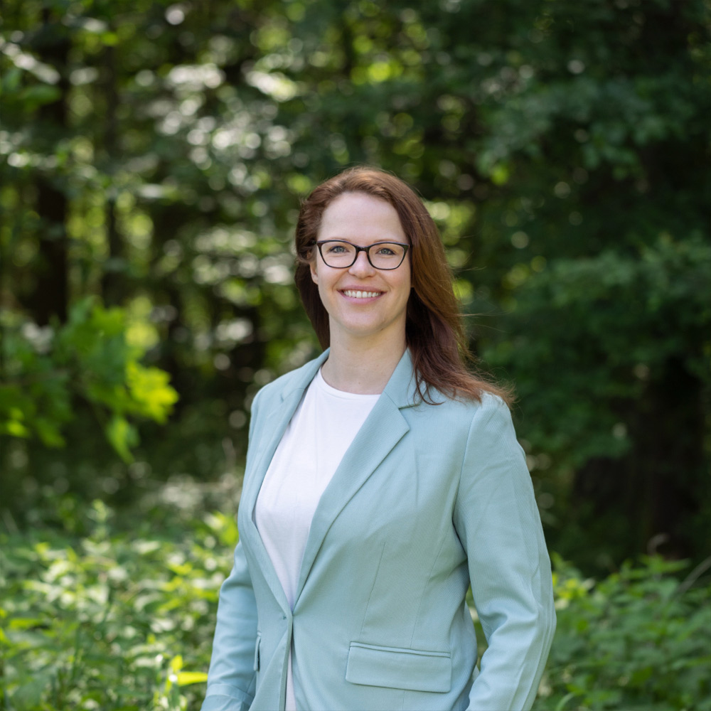 Anne Kayser lächelnd im türkisfarbenen Blazer, stehend vor Bäumen im Wald. Sie schaut direkt in die Kamera und repräsentiert ihre Rolle bei VitaPart® als analytische und kreative Kraft, die sowohl für die rechtliche Sicherheit der Produkte als auch für ihre ansprechende Präsentation sorgt.