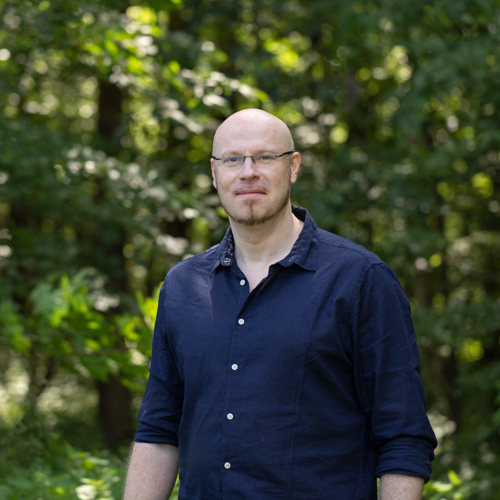 Dr. Steffen Jakobs lächelnd im blauen Hemd vor Bäumen im Wald. Er schaut direkt in die Kamera und repräsentiert seine Leidenschaft für die Produktentwicklung bei VitaPart®, bei der er sich auf ernährungswissenschaftliche und medizinische Studien stützt, um hochwertige Präparate zu schaffen.